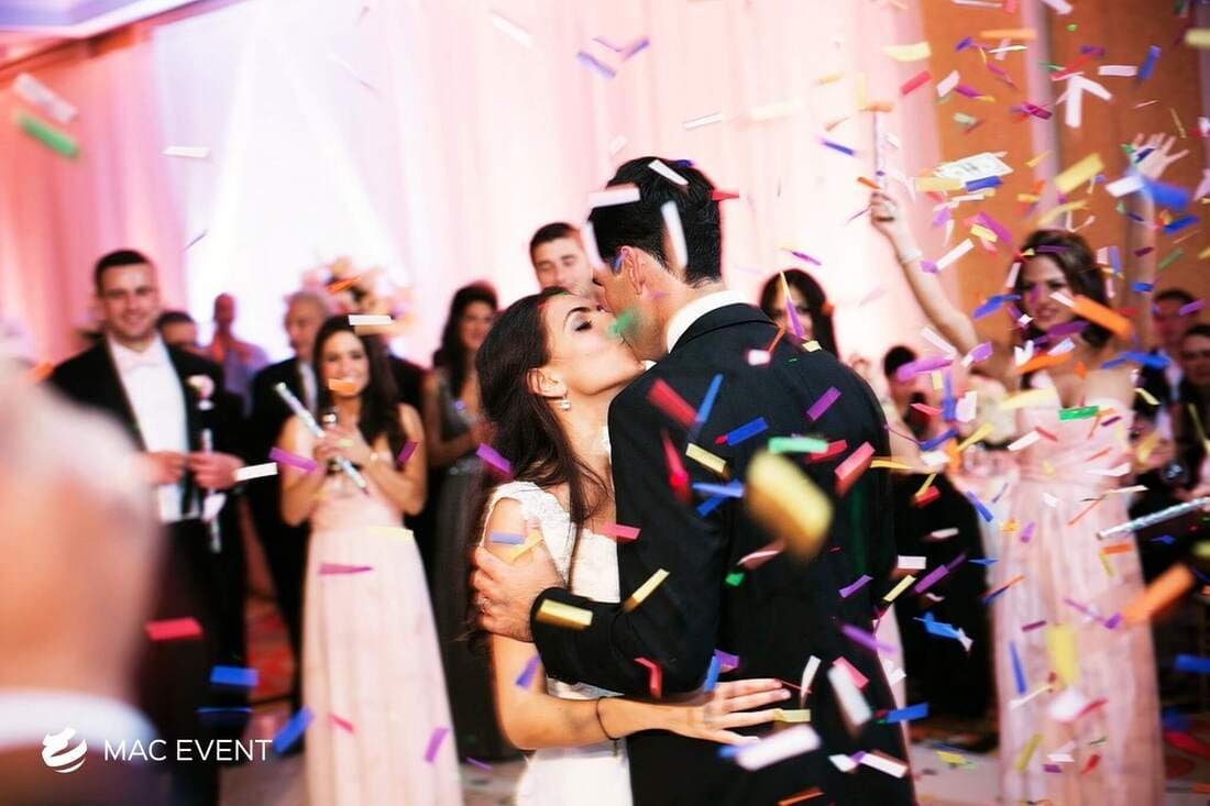 A bride and groom share a romantic kiss on the dance floor as colorful confetti rains down, surrounded by wedding guests in an elegant ballroom setting.