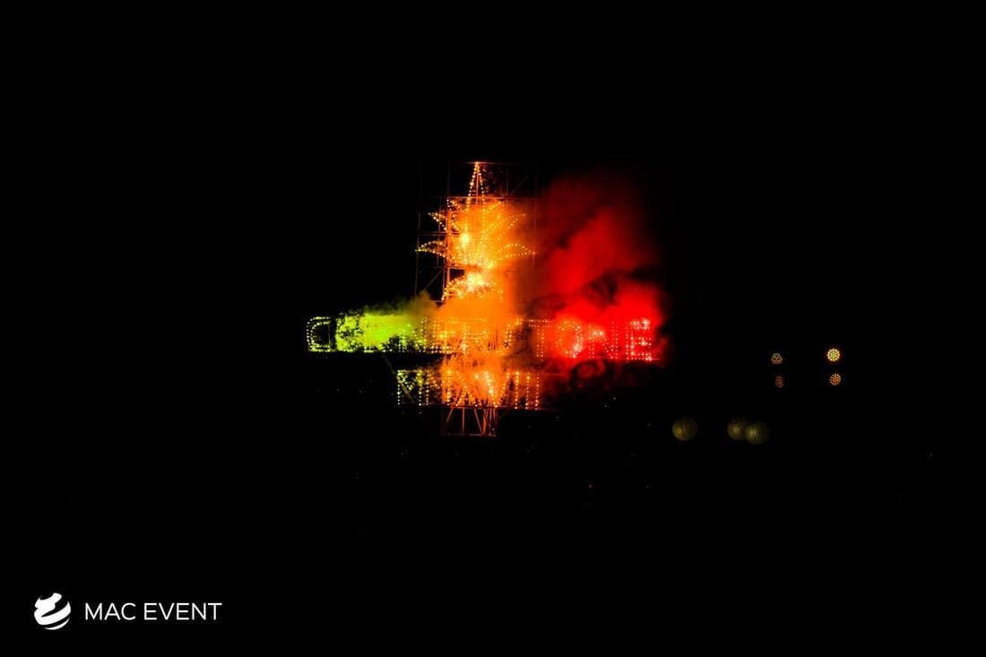 A dramatic firework display with multicolored lettering and a bright star-shaped explosion, partially obscured by smoke, creating an intense visual impact at an event.